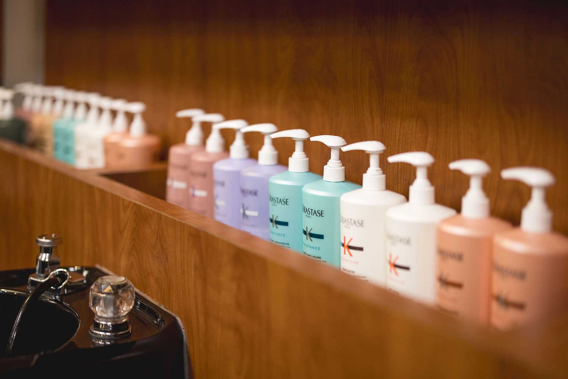 A row of various colorful shampoo and conditioner bottles lined up on a wooden shelf in a salon.