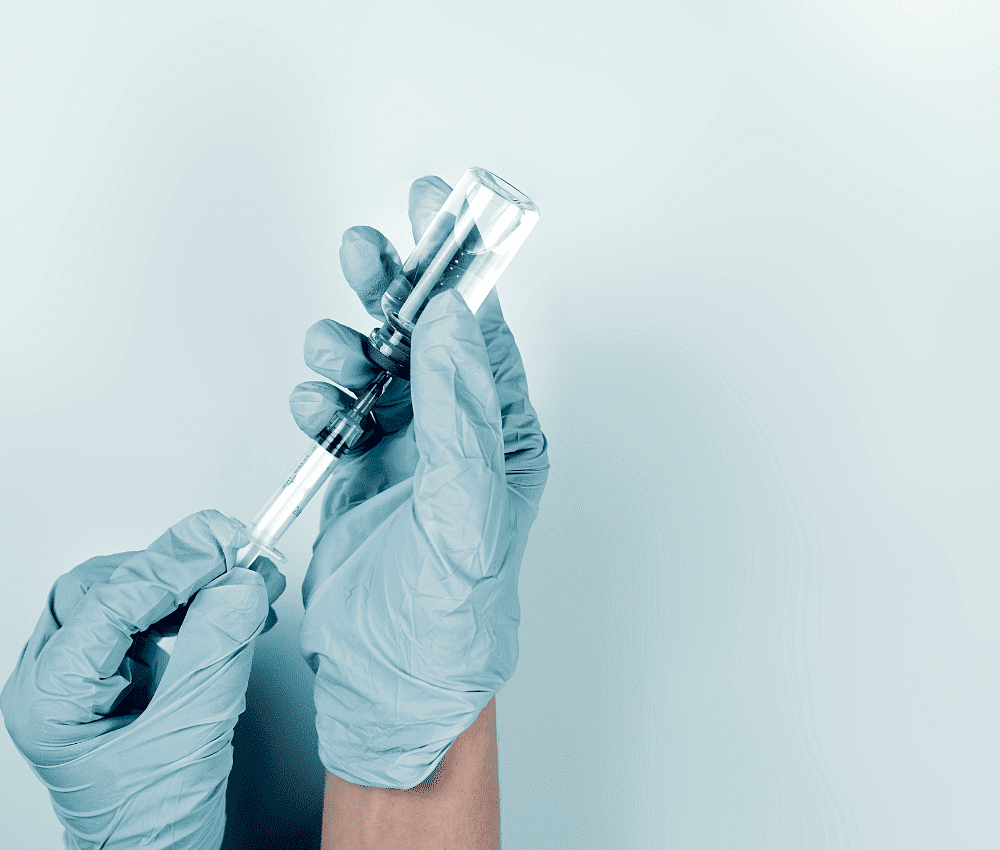 Person in gloves filling syringe from vial against blue background.